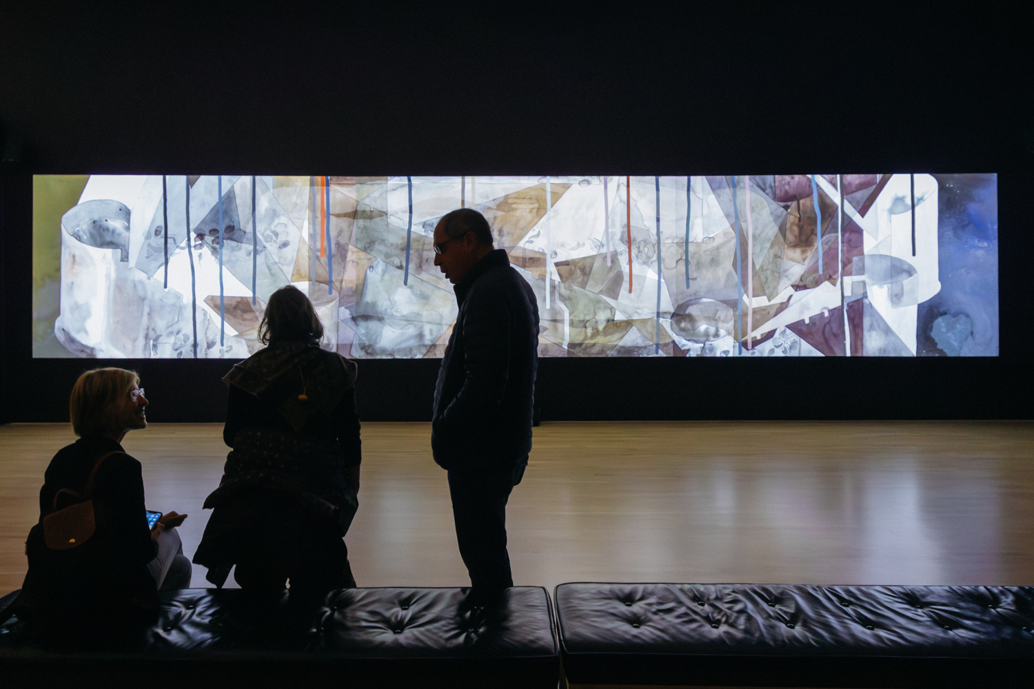 A large, wide screen in a dark room depicts abstract imagery. Two viewers are seated on a couch in the foreground, while another stands nearby, observing the artwork.