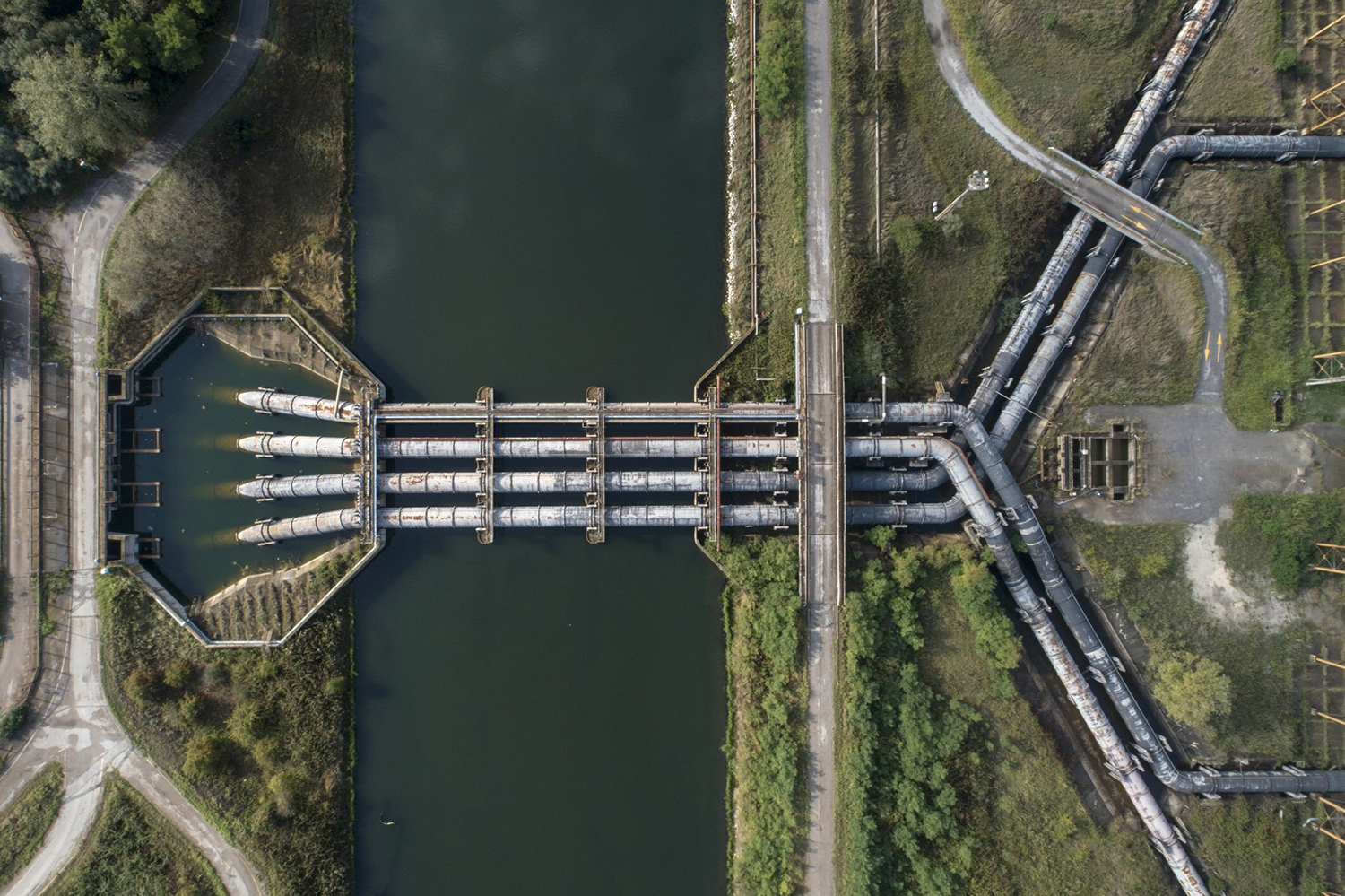 Aerial view of an industrial waterway with multiple large pipelines crossing over a river surrounded by greenery.