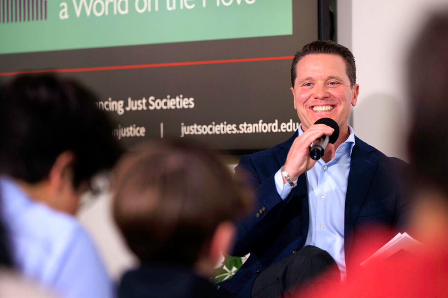Tomás Jimenéz engages an audience during a discussion about advancing just societies, with a display board featuring relevant information in the background.