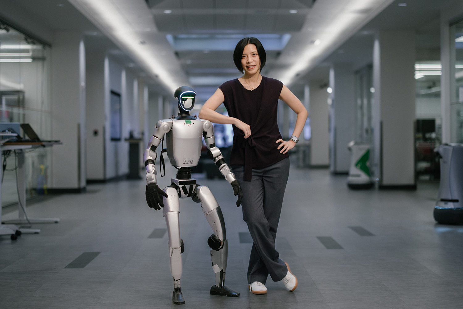 Karen Liu stands beside a humanoid robot in a modern indoor setting, both showcasing a friendly pose.