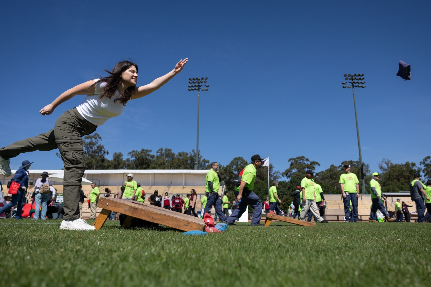 A person throwing a bean bag toward a target during a community event, with many people in bright green shirts in the background.