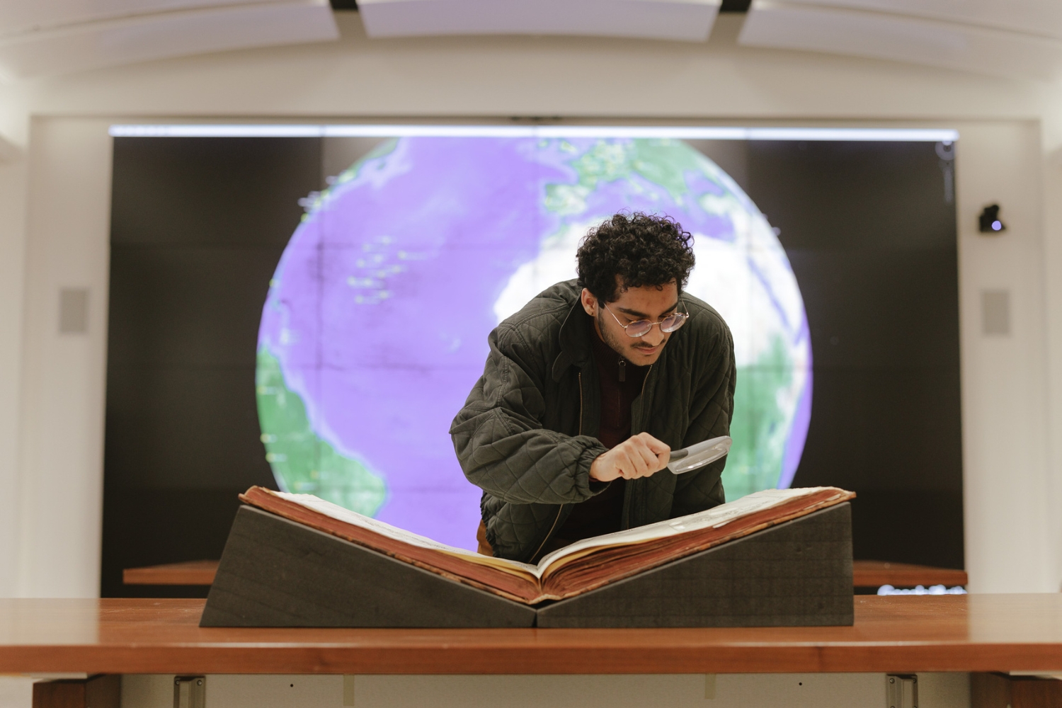 A person examines an open book with a magnifying glass in front of a large globe display.
