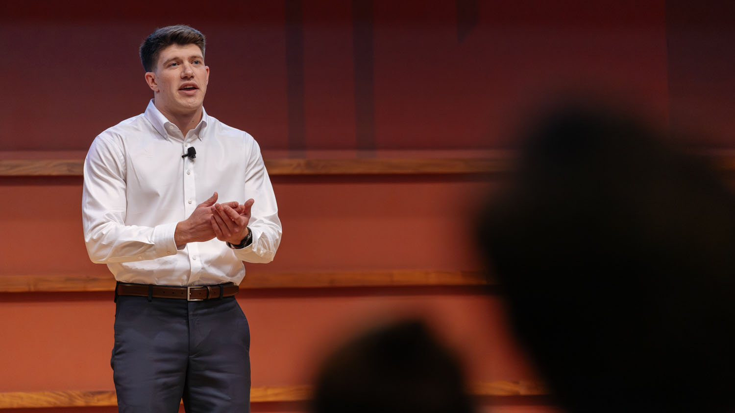 AJ Phillips in white shirt speaks to audience on stage at 3MT competition.