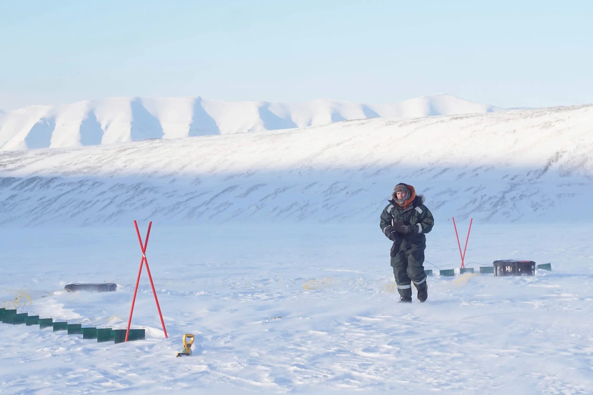 A person in a heavy winter jacket walks through a snowy landscape, with mountains and markers visible in the background.