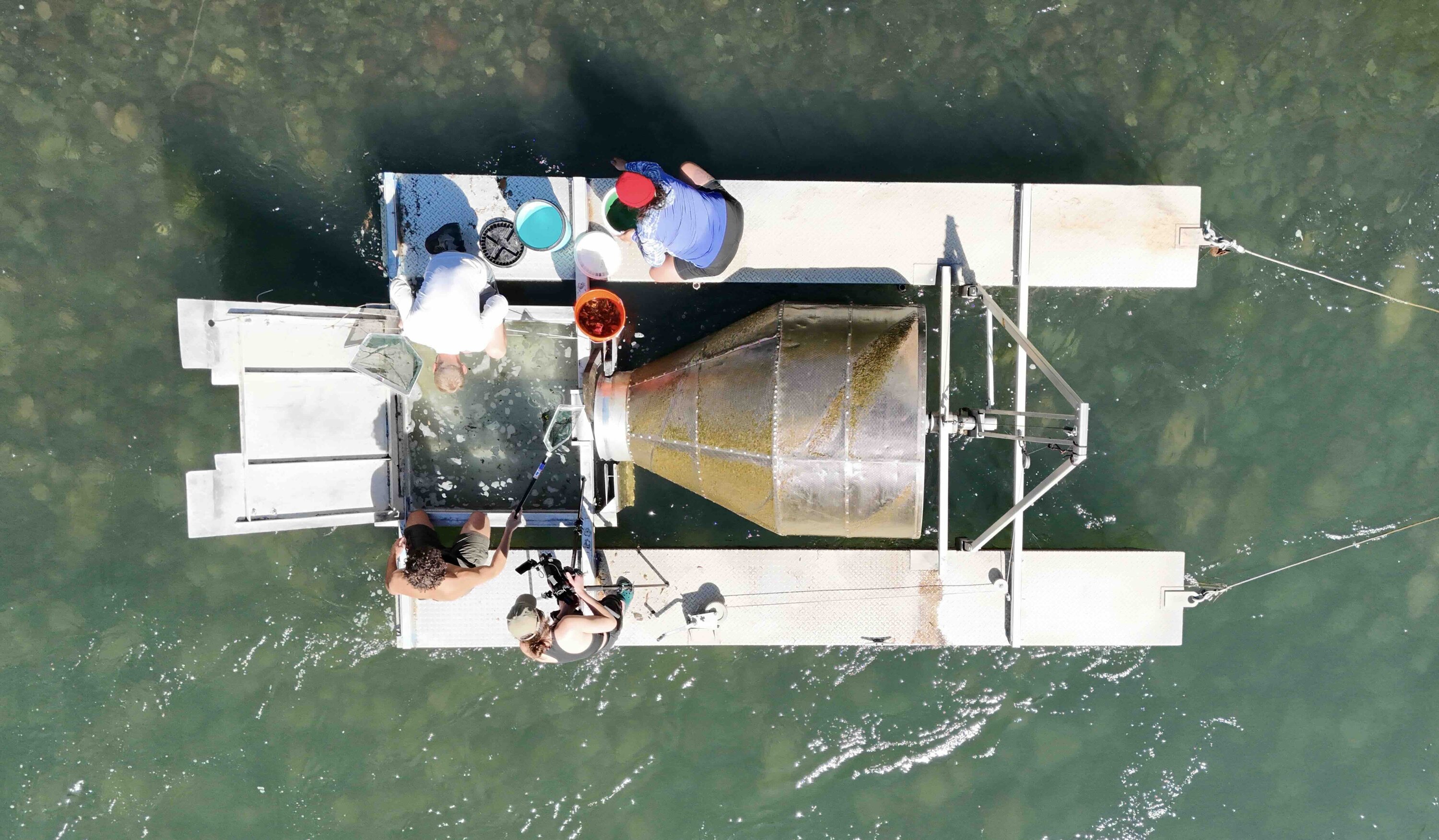 Researchers work on a floating platform in the water, collecting samples using nets and buckets near a large cylindrical device.