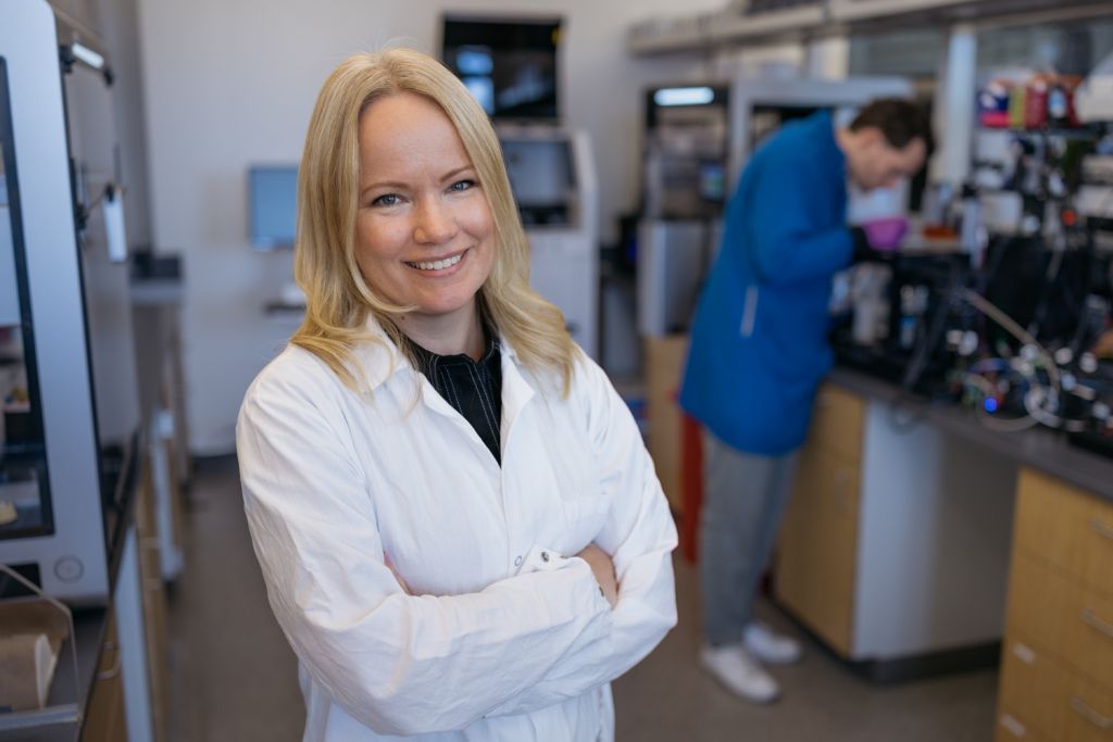 Emma Lundberg in a lab coat standing and posing for a photo, while a colleague examines equipment in the background.