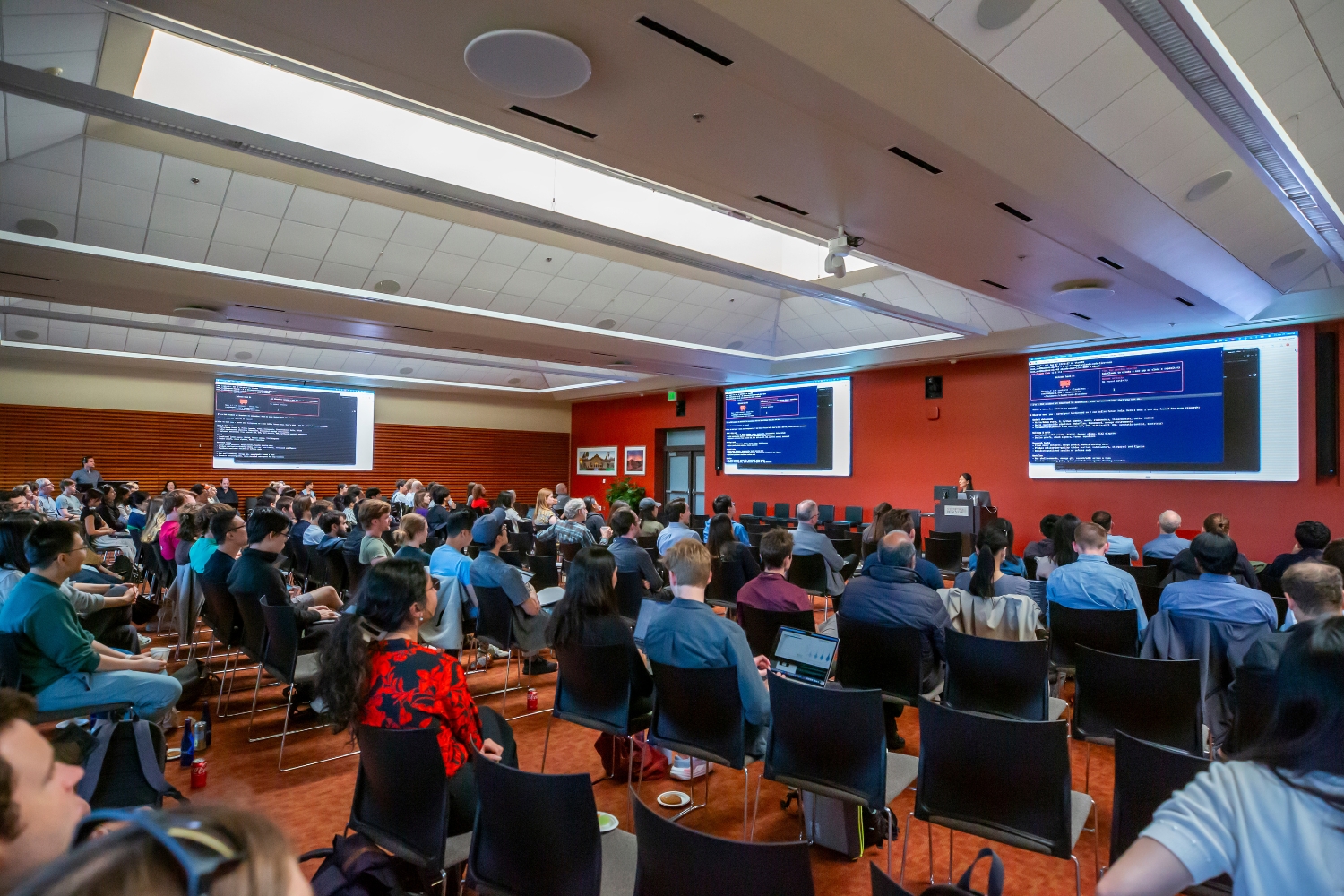 An audience listens to a speaker giving a demonstration on large screens.