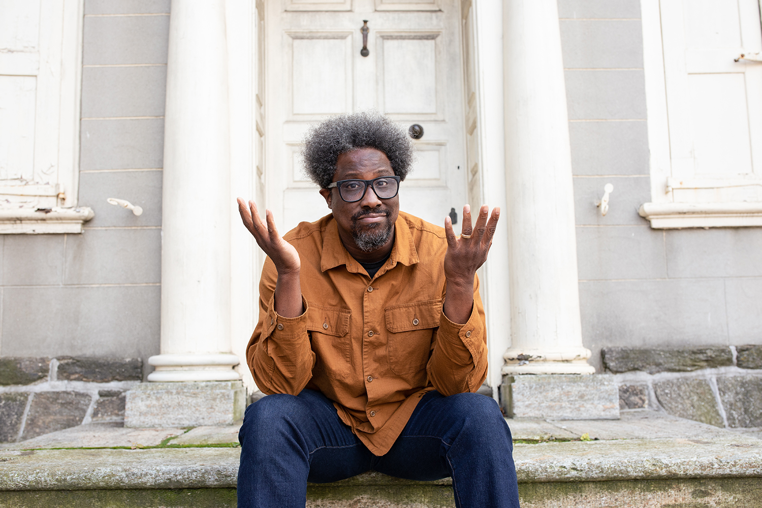 Bell sits on steps, raising his hands expressively as if questioning or reflecting, in front of an old building.