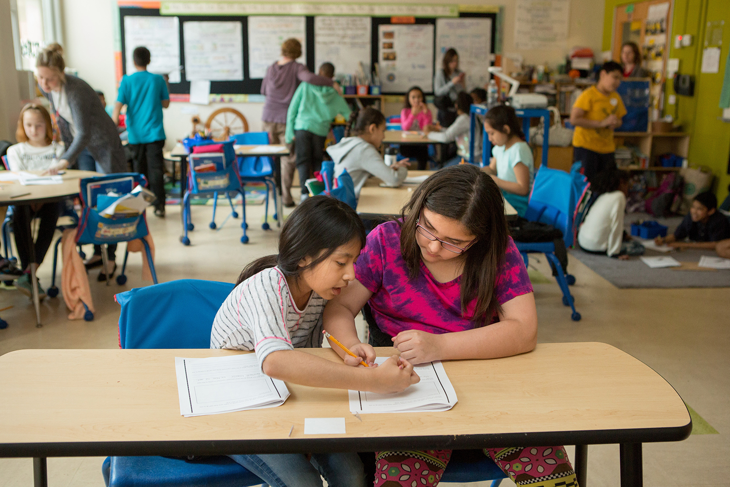 Two girls collaborate on assignments at a classroom table, while other students engage in various activities in the background.