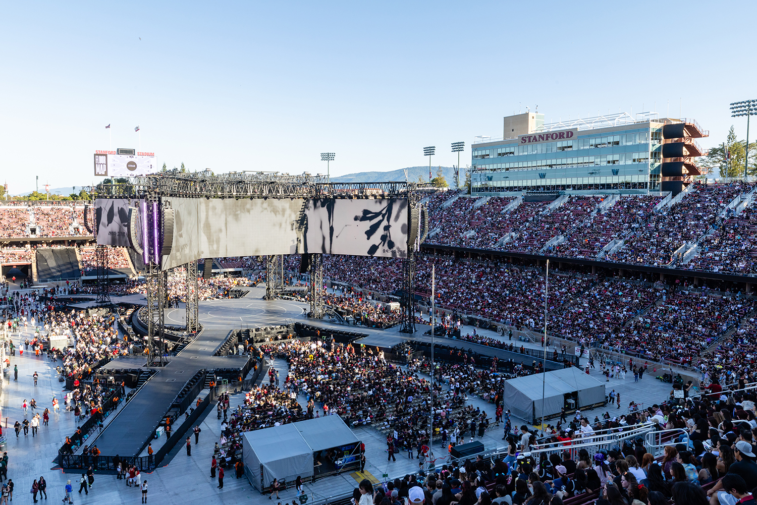 A packed stadium filled with fans anticipates a performance, showcasing a large stage and video screen under a clear sky.
