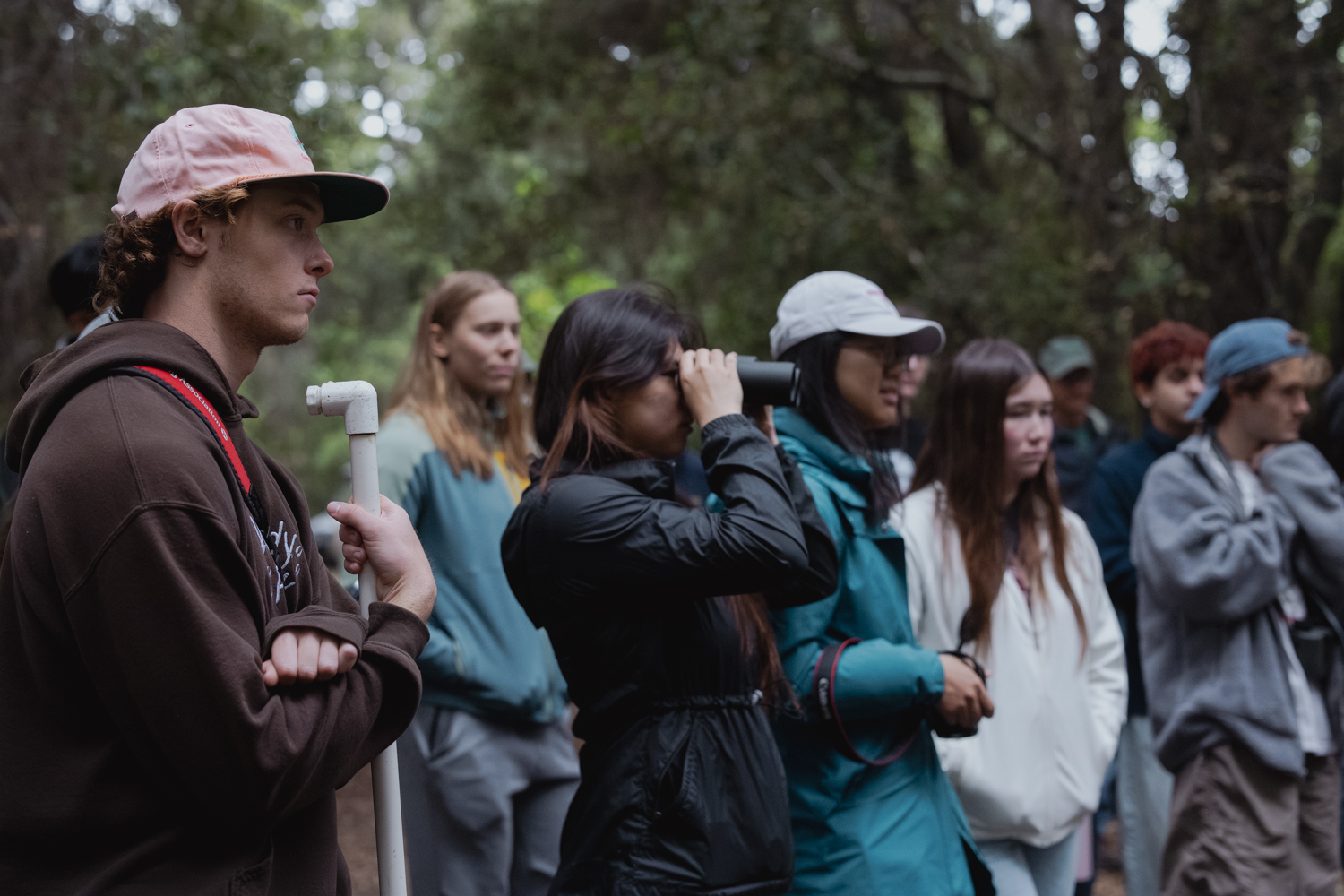 With a wooded area in background, students , one with binoculars,are gathered looking toward the right.