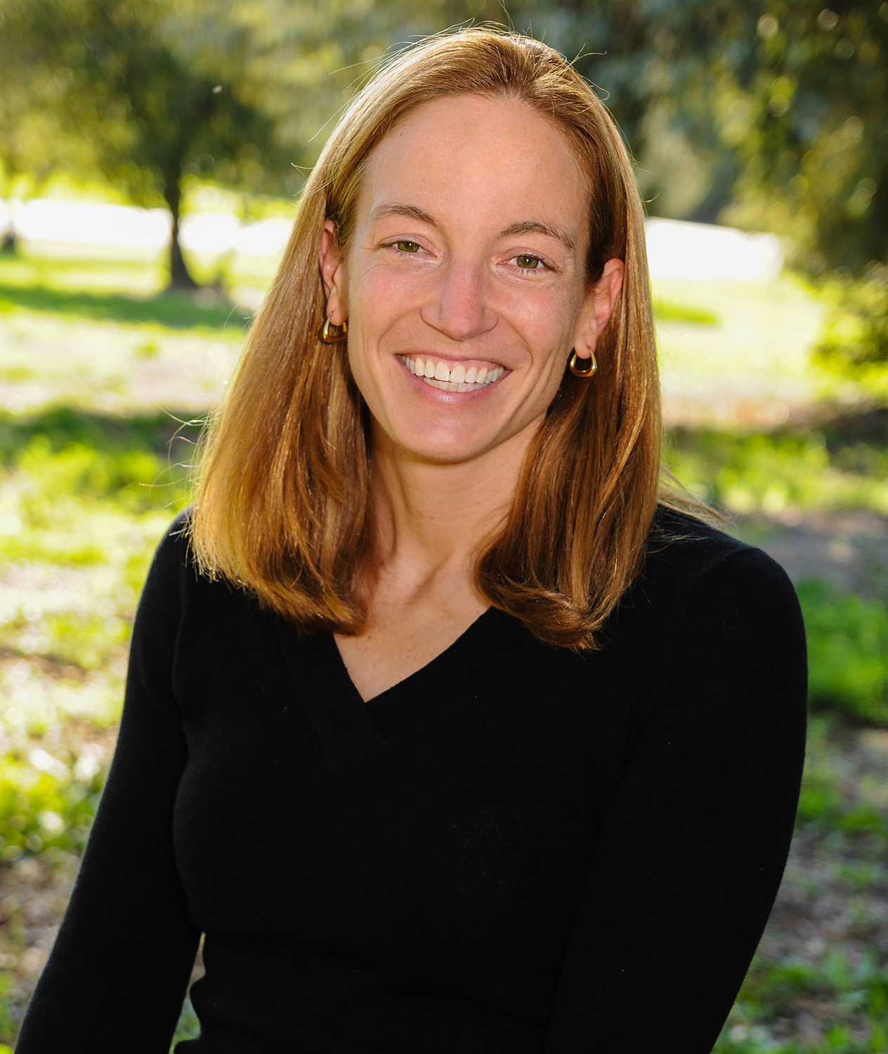 Salmeen with long reddish-brown hair smiling outdoors in a park-like setting.