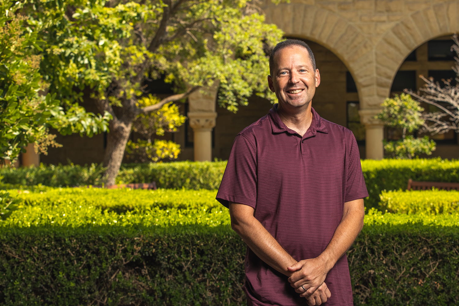 Ira Lit in a maroon polo shirts stands in a garden with green hedges and trees edging the Quad arcades.