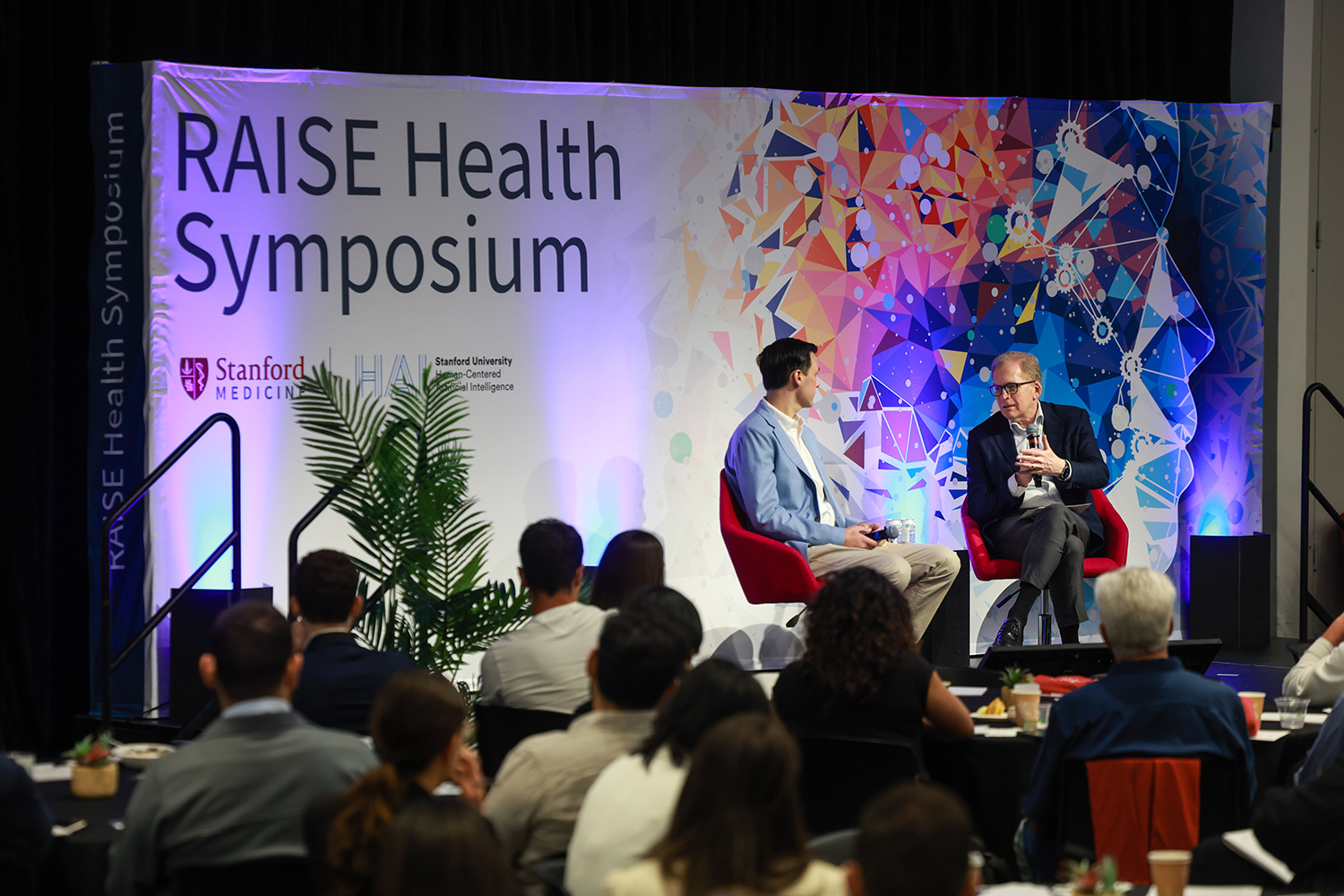 Two speakers discuss topics on stage at the RAISE Health Symposium.