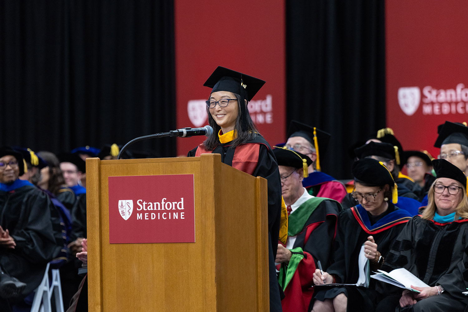 Graduate in cap and gown smiling while speaking at podium during Stanford Medicine commencement ceremony.