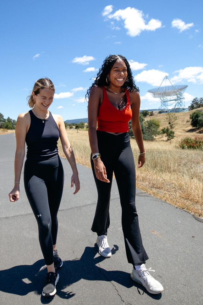 Two friends walk together while laughing at their conversation while on a sunny path, with a large radio telescope visible in the background.