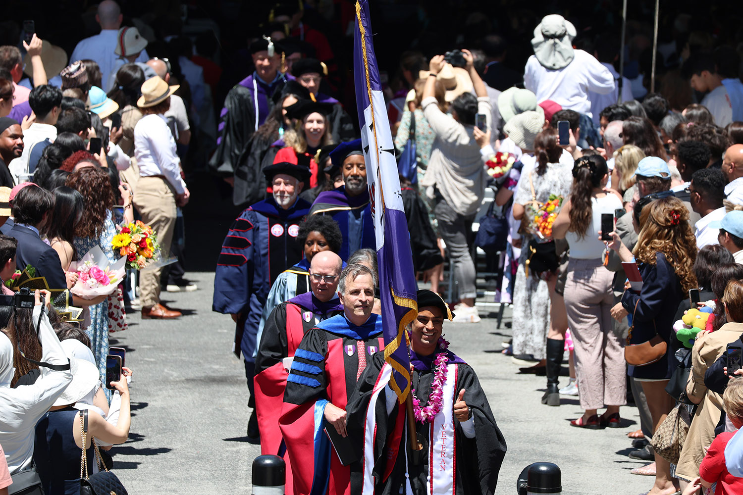 Procession in academic regalia, while family and friends cheer and take photos from the sidelines.