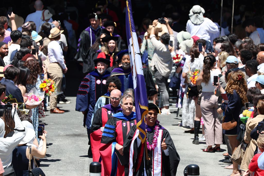 Procession in academic regalia, while family and friends cheer and take photos from the sidelines.