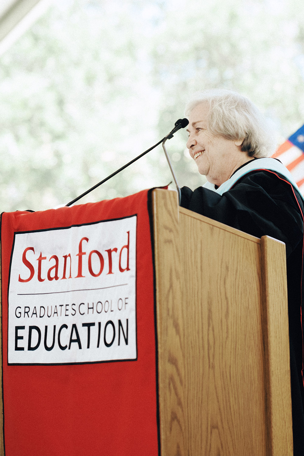 Speaker Rachel Lotan wearing academic robes addresses graduates at a podium displaying the Stanford Graduate School of Education banner.