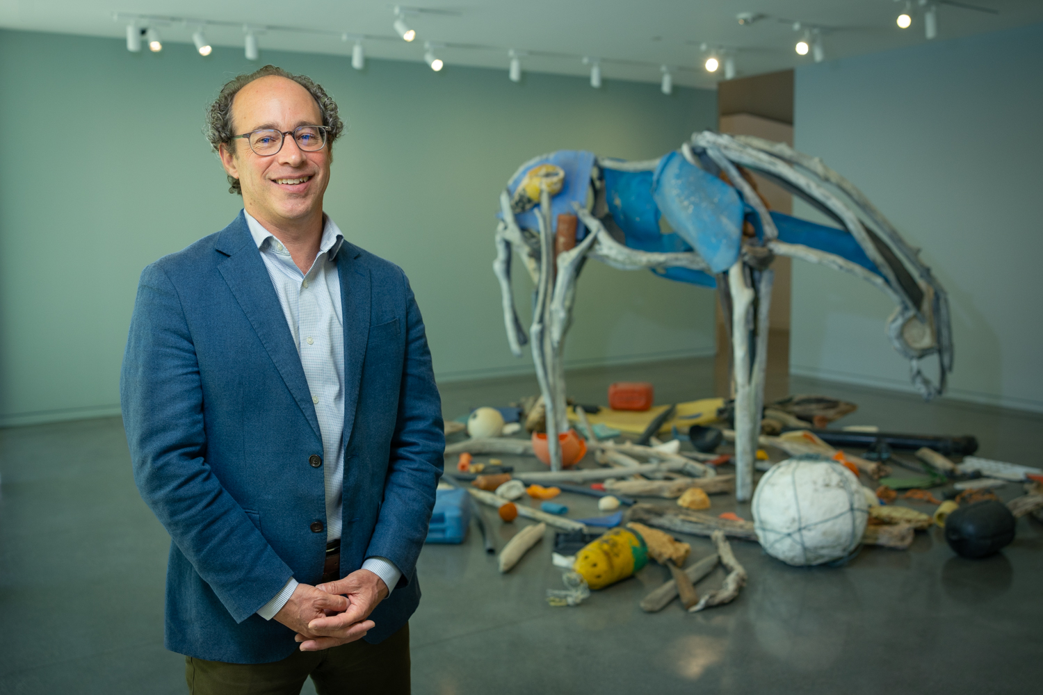 Linetzky stands beside a large horse sculpture made of various materials, including colored objects and driftwood, in art gallery.