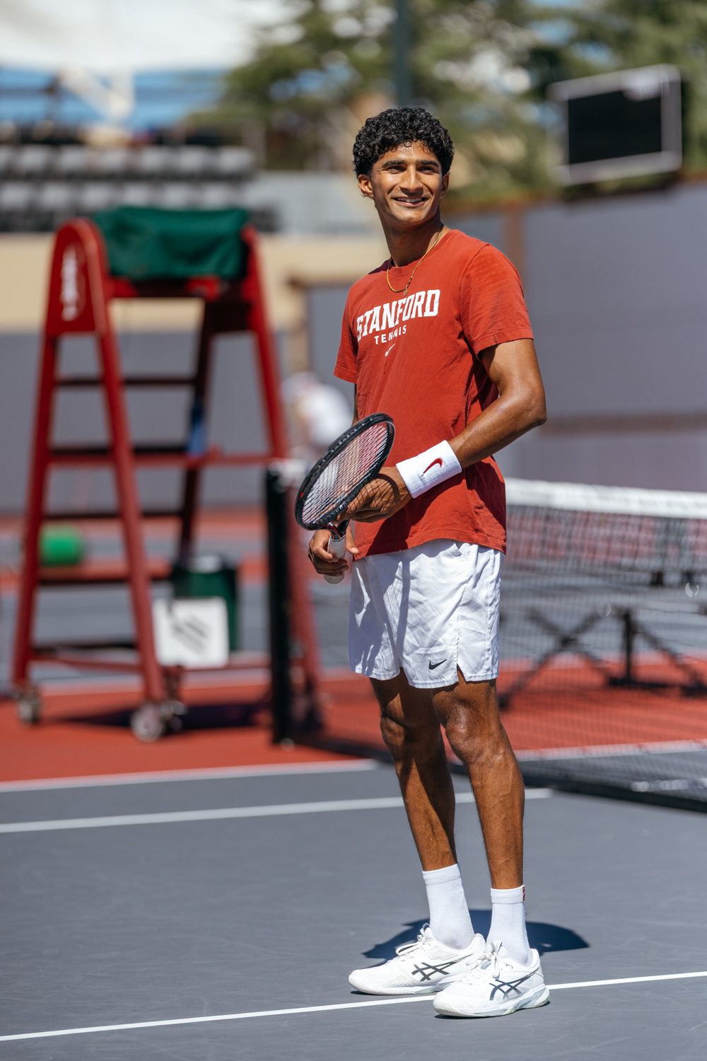 Samir Banerjee smiles while holding a tennis racket on an outdoor court, wearing a red Stanford tennis shirt and white shorts.