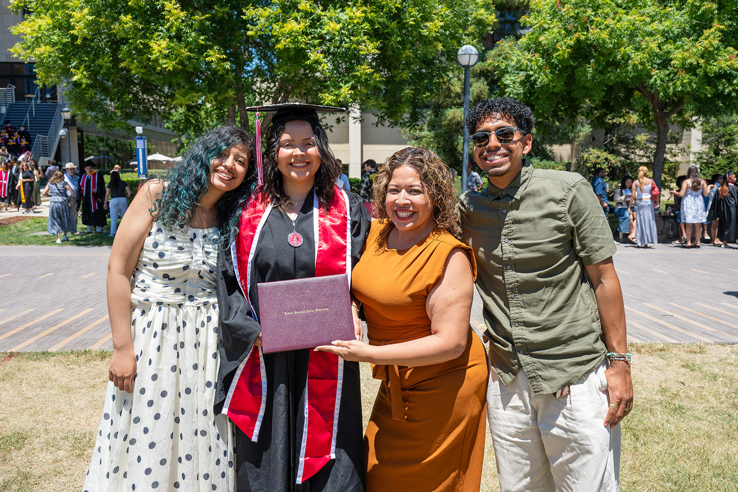 Graduate holds a diploma while posing with three family members outdoors at a graduation celebration.