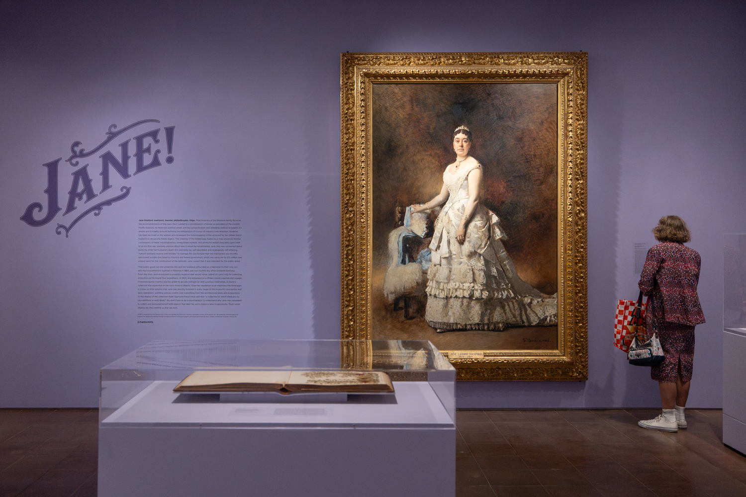 A visitor admires a large portrait of Jane Stanford in an ornate dress, displayed in an exhibition titled 