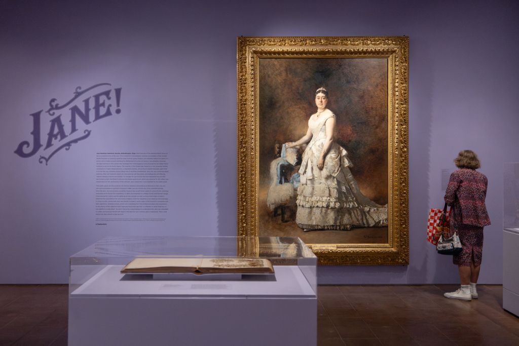 A visitor admires a large portrait of Jane Stanford in an ornate dress, displayed in an exhibition titled 