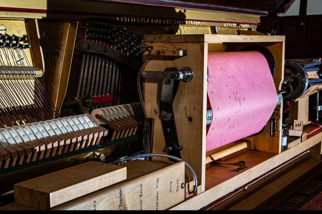 A player piano's mechanism holds a pink piano roll on its spool, with hammers and strings visible inside the open piano case.