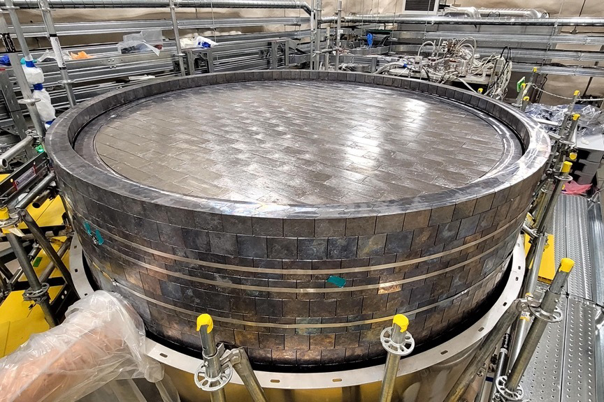 A large cylindrical structure built from stacked dark metal bricks sits on a circular platform inside a laboratory hall.