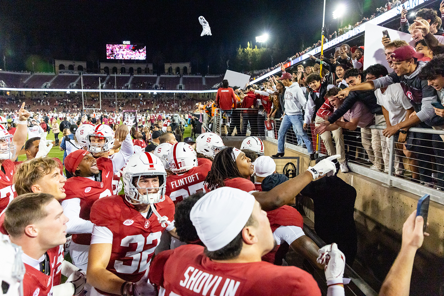 Cardinal players celebrate with fans along the stadium railing after a game, as someone tosses a mascot head into the air.