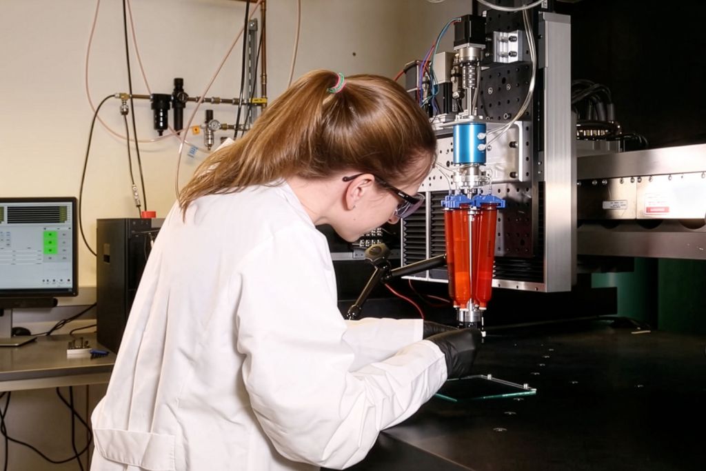 A researcher in a lab coat operates a 3D printing device that deposits red material through a nozzle onto a glass plate.