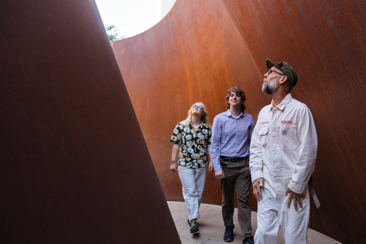Two people and guide Mark Shunney in white coveralls walk between the leaning rust-colored steel walls of a curving sculpture.
