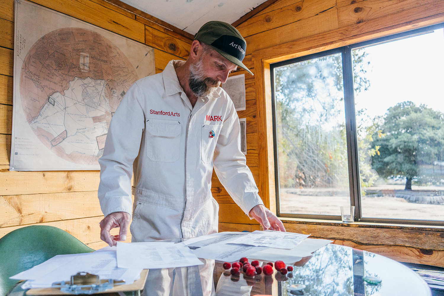 Mark Shunney in a white Stanford Arts coverall and cap reviews printed documents on a glass table in a wood-paneled room.