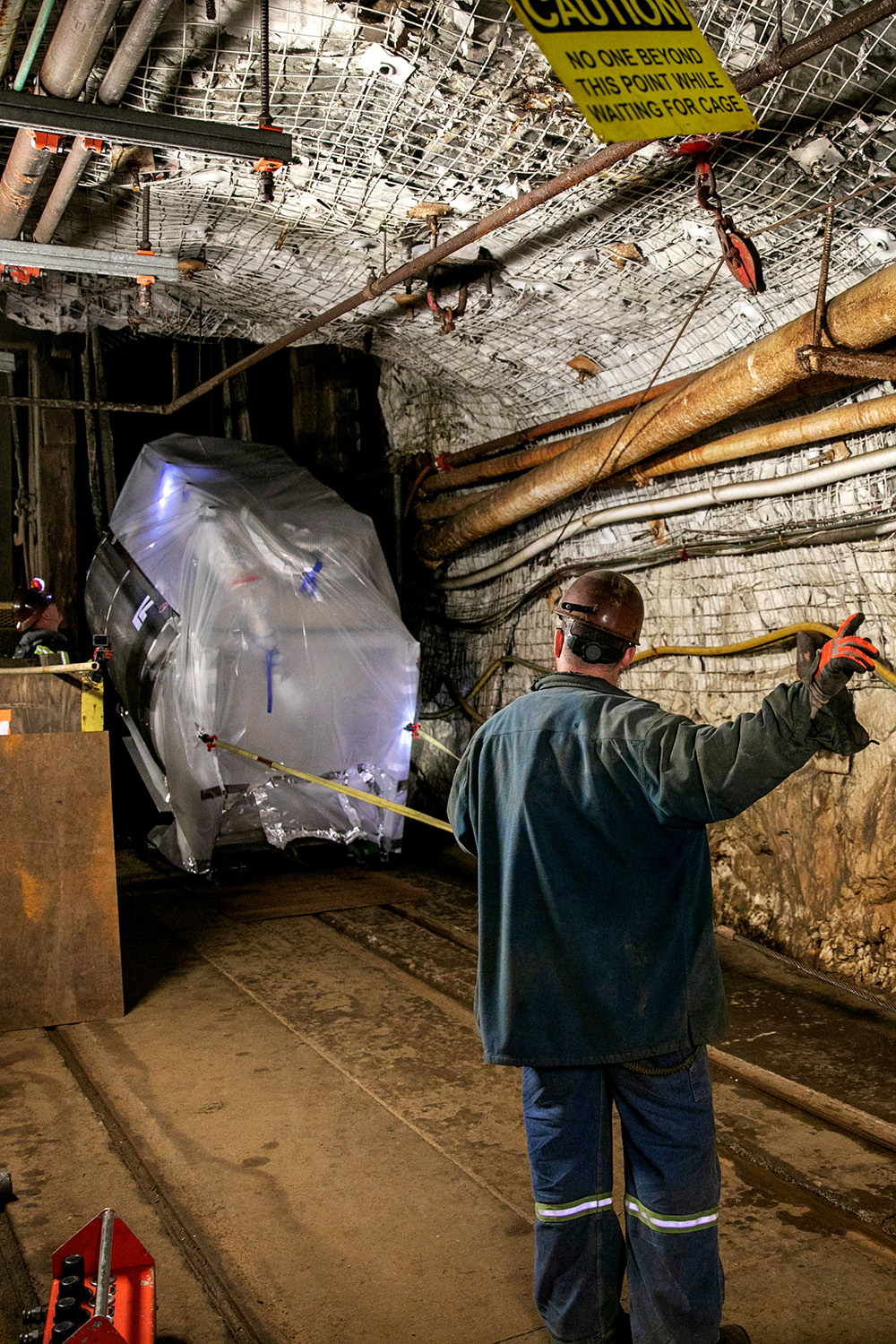 A worker in a hard hat guides a large plastic-wrapped detector through an underground mine tunnel.