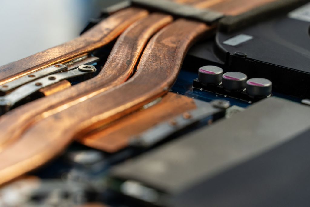 Macro shot of copper heat pipes on a laptop's circuit board, with capacitors and screws visible, shallow depth of field.