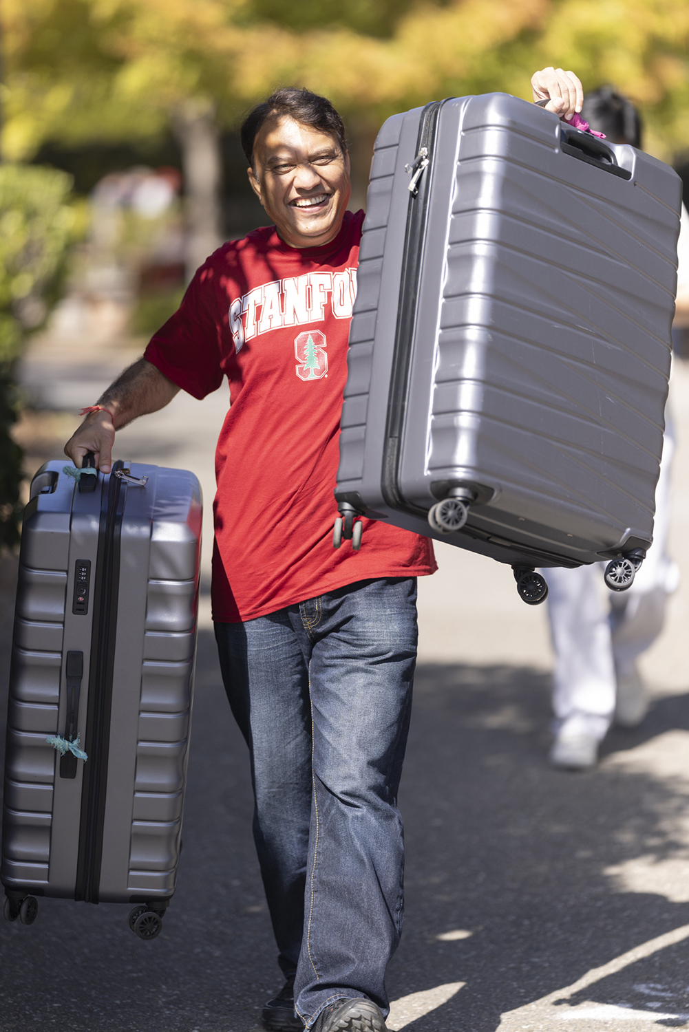A father proudly carries two suitcases while walking toward his student's dorm on a tree-lined street.