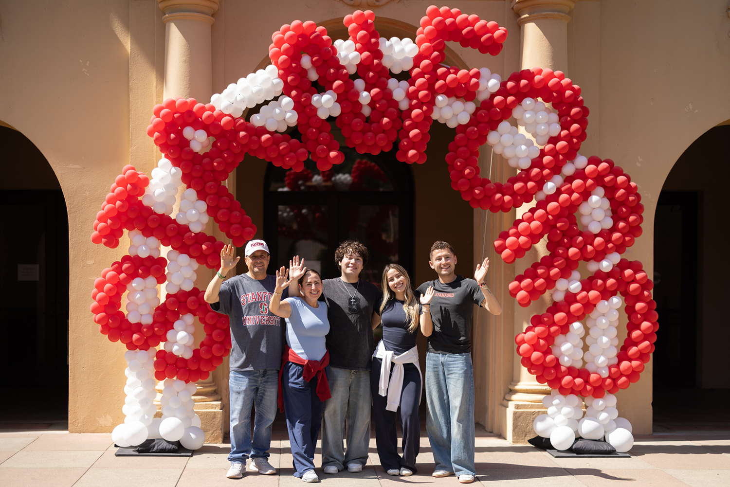 A family poses together beneath a large red and white balloon arch outside a campus building.