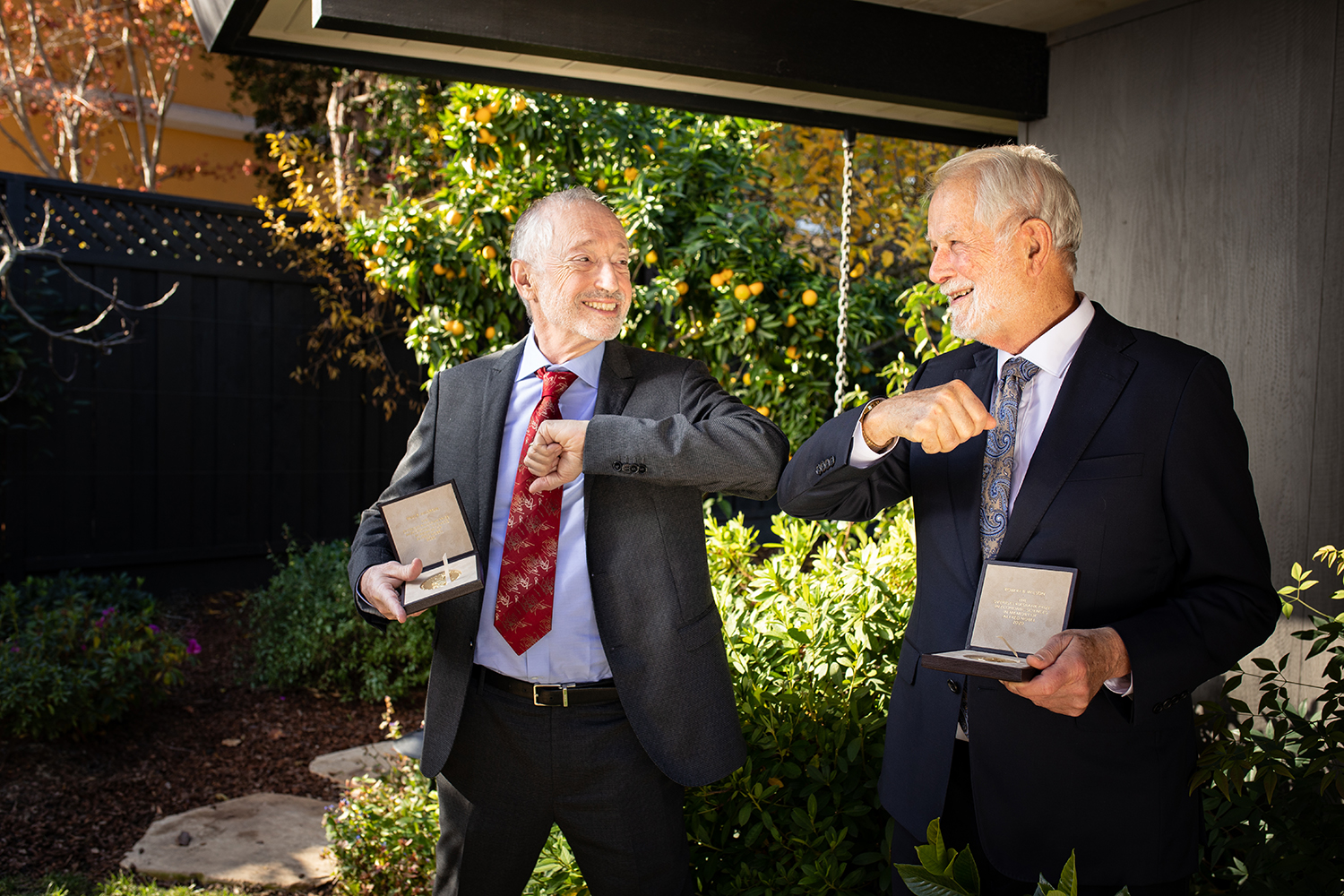 Paul R. Milgrom and Robert B. Wilson bump elbows in a garden, each holding an open Nobel medal case.