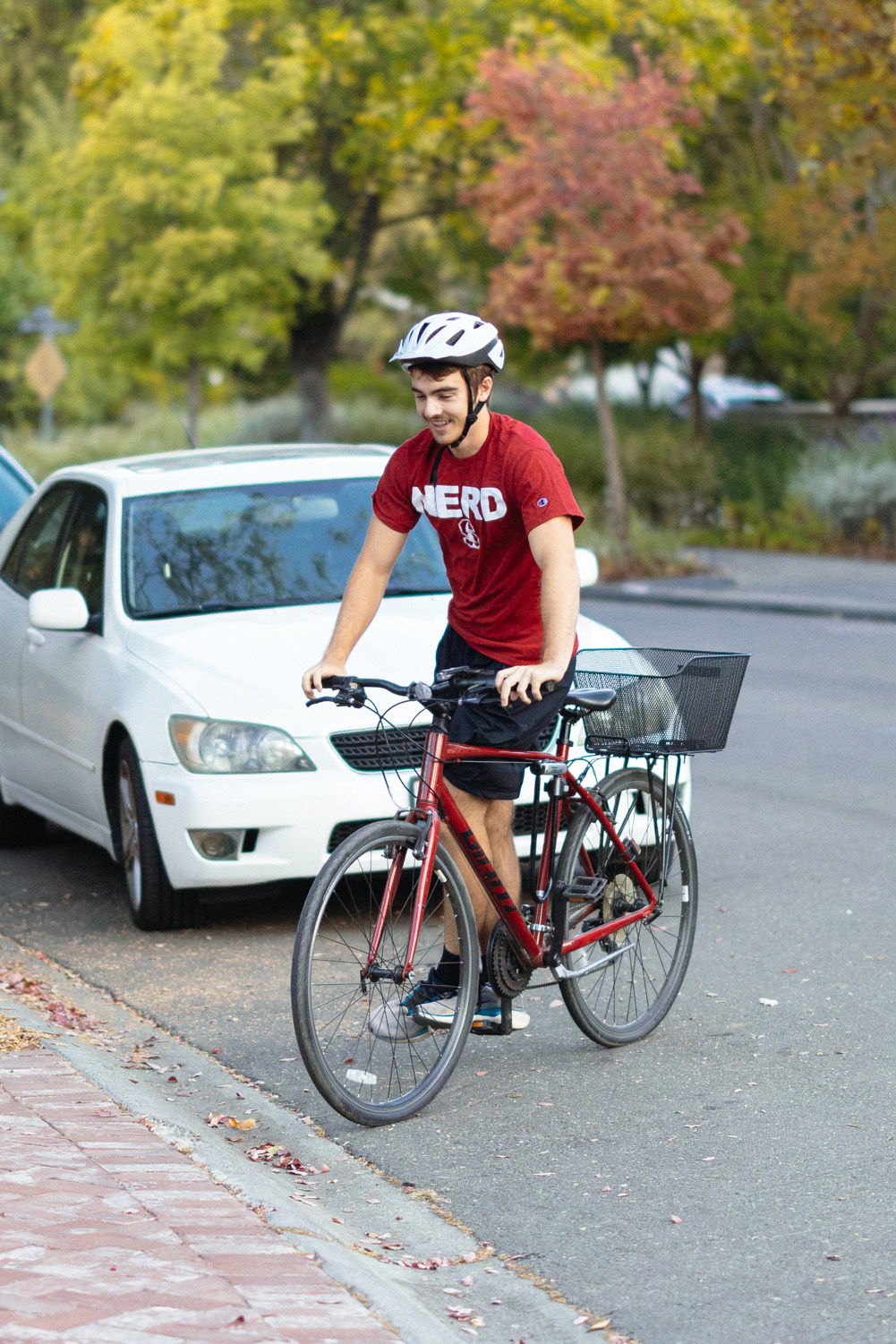 Alexander Deisseroth red Stanford NERD T-shirt and white helmet walks his bike along a tree-lined street.