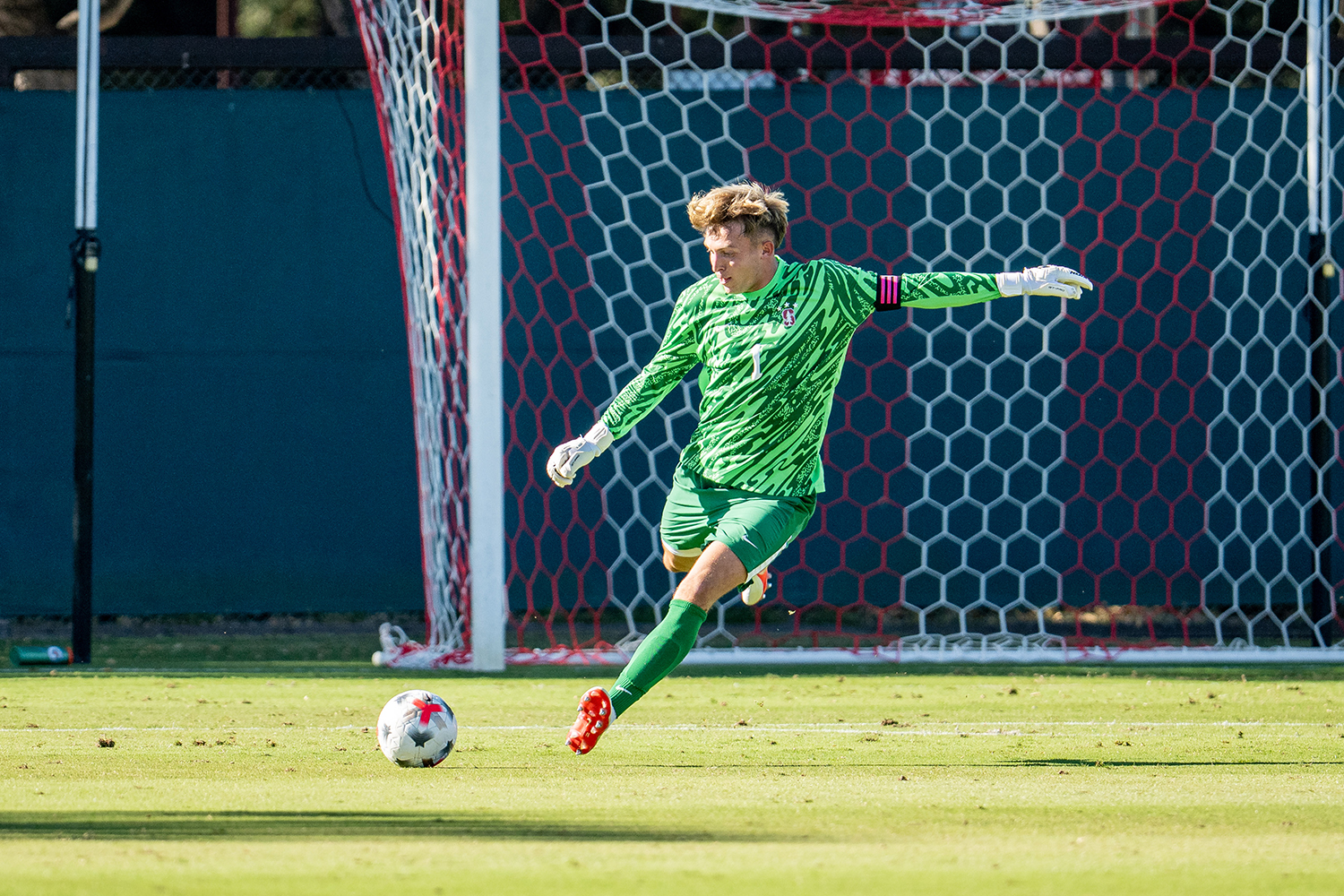 A Cardinal goalkeeper winds up to kick a soccer ball in front of the goal net.