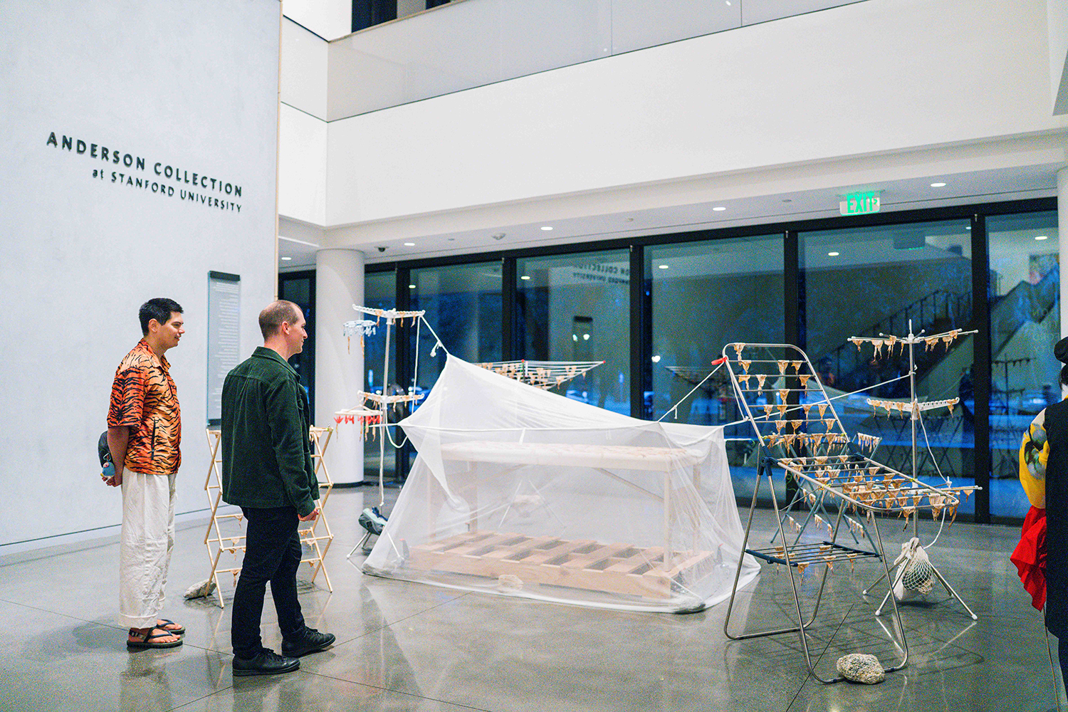 Two men look at an installation of drying racks and a net-covered table in a museum lobby.