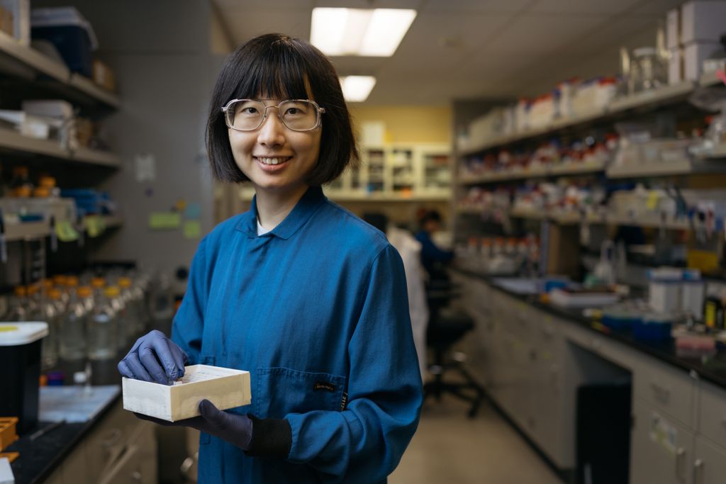 Nina Zhao in safety glasses and a blue lab coat smiles while holding a box of samples in a lab aisle lined with shelves of supplies.