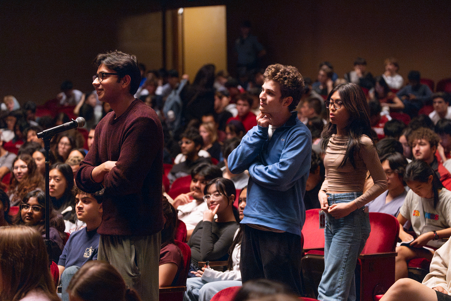 A student stands and speaks into a microphone in a crowded auditorium, with two other students lined up behind him during a Q&A portion of a panel event.
