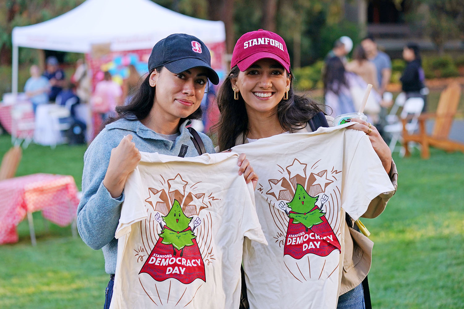 Two smiling students in Stanford caps hold up matching event T-shirts on a lawn with tents and picnic tables in the background.