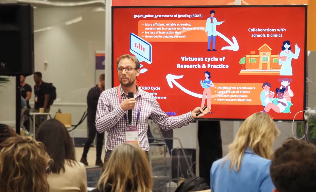 Professor Jason Yeatman speaks to a group in front of a red slide depicting the "virtuous cycle of research and practice" of ROAR.