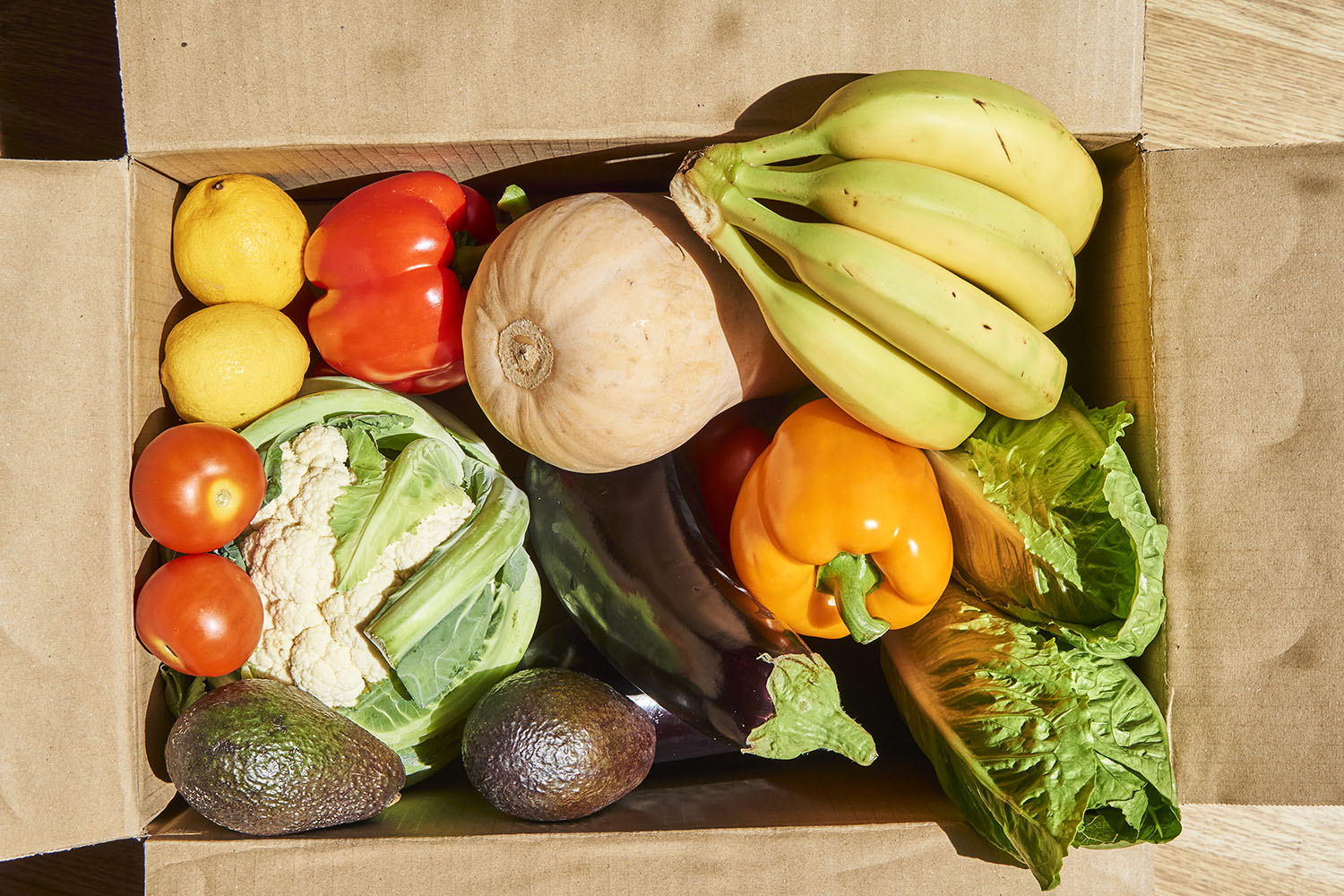 Cardboard box containing produce including cauliflower, butternut squash, bell peppers, avocadoes, bananas, and more