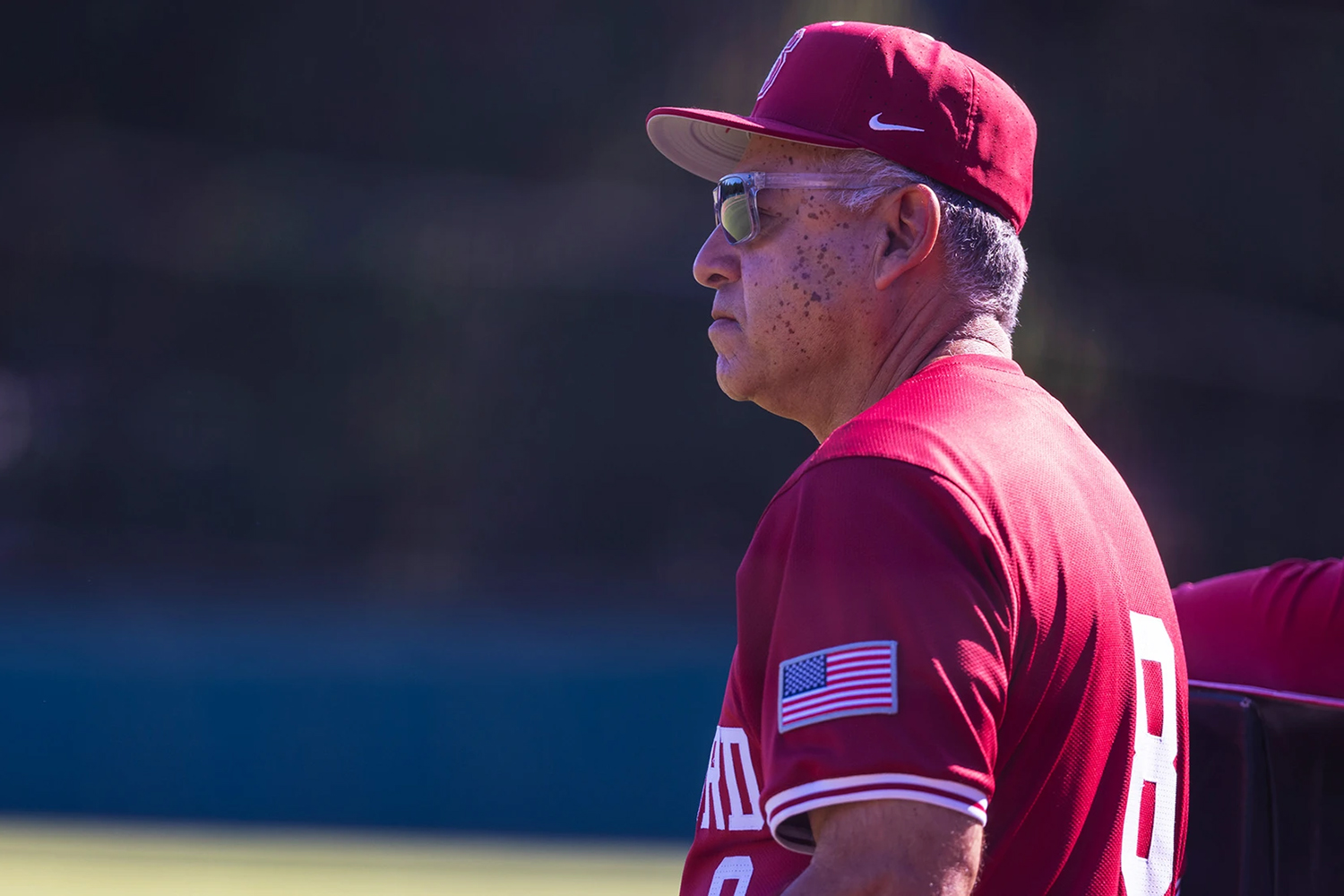 Baseball coach David Esquer in a red jersey with an American flag patch on the sleeve watches a game, standing against a green backdrop in bright sunlight.