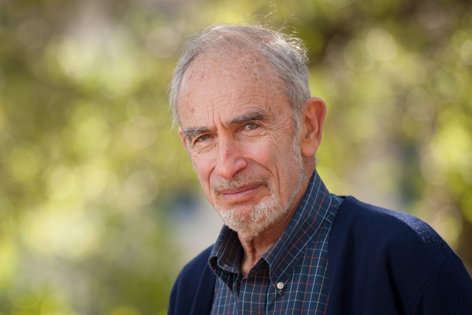 Paul Ehrlich posing for a photo outdoors, wearing a plaid shirt and dark sweater, surrounded by a soft, blurred green background.