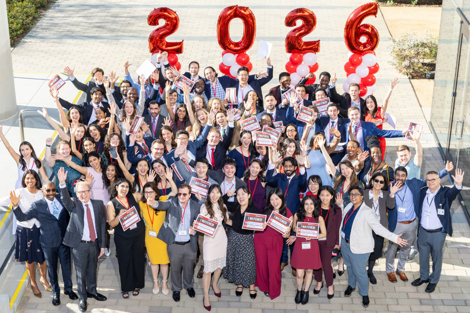 A large group of smiling individuals poses with signs and giant balloons, celebrating Match Day for the Class of 2026.